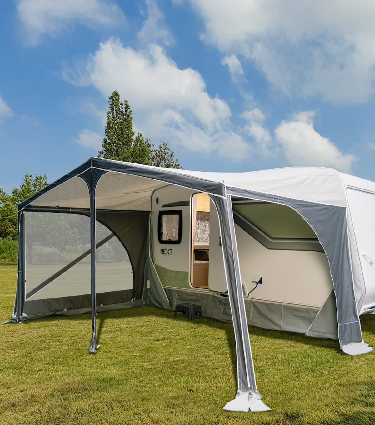 Weißer NEXT Caravan mit großem, grauem Vorzelt auf einer grünen Wiese unter blauem Himmel beim komfortablen Camping